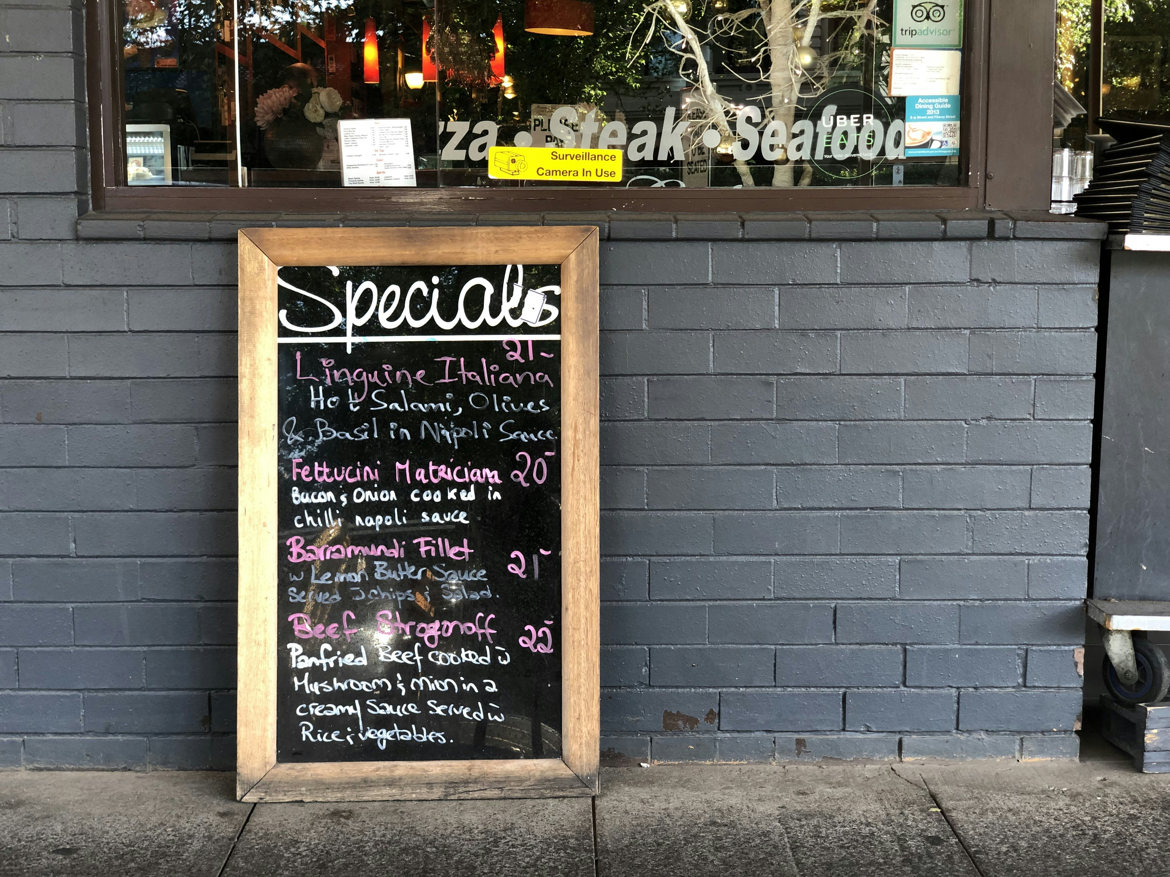 A restaurant exterior features a menu board with handwritten specials in various colors. The board lists dishes such as Linguine Italiana, Fettuccini Mataciana, Barramundi Fillet, and Beef Stroganoff with their respective prices. It leans against a gray brick wall, and part of the restaurant's name is visible through the window behind, alongside some seating and menus displayed inside.